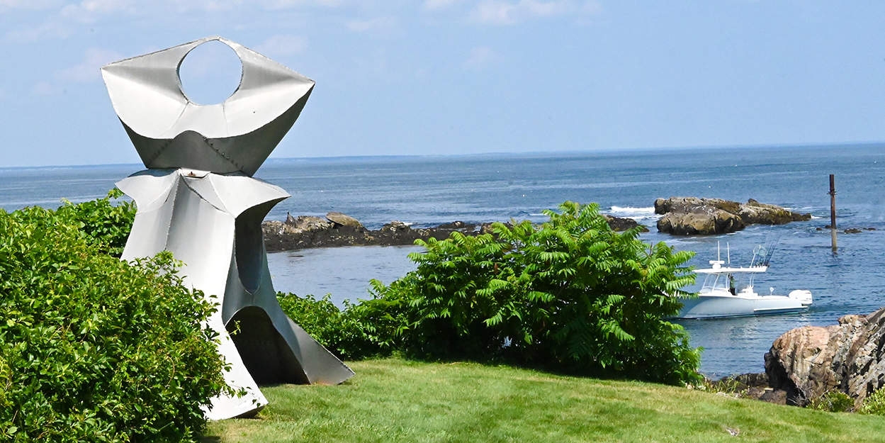 a tin sculpture on a lawn and the ocean and boat in the background
